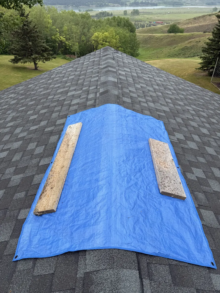 Blue emergency tarp weighted with planks over storm-damaged shingles below the ridge of a Calgary roof